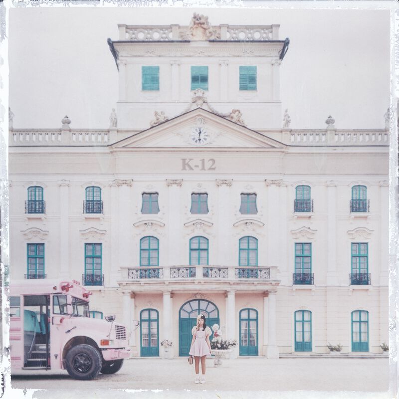 The cover of Melanie Martinez's album titled K-12. We can see her standing in front of a school looking building on the front of which it is written